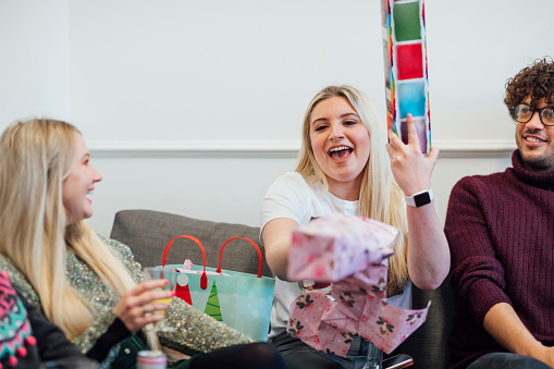 Office staff enjoying a Christmas party together, unwrapping gifts as part of secret Santa. A woman is holding up a gift while laughing. They are in the North east of England.