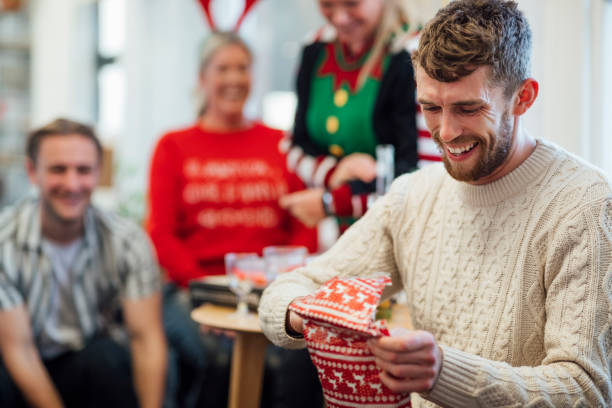 Unwrapping a Christmas Present Office staff enjoying a Christmas party together, unwrapping gifts as part of secret Santa. A man is unwrapping a gift while laughing. They are in the North east of England. office christmas party stock pictures, royalty-free photos & images