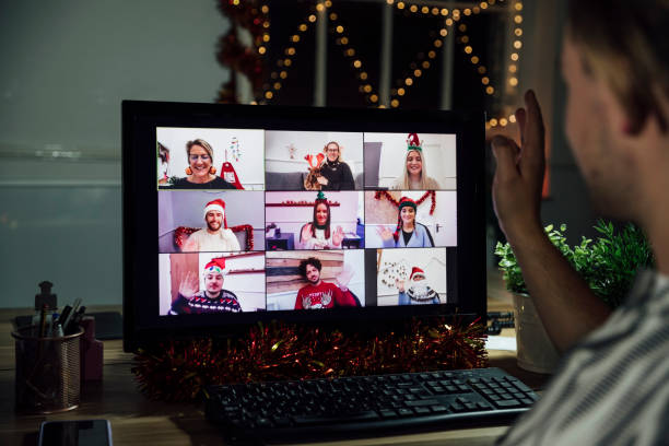 Celebrating Safely Over the shoulder view of a festive group of colleagues having an office party via video call. They are all greeting each other and chatting whilst wearing festive clothing. A man is sitting at his computer having to the screen. office parties stock pictures, royalty-free photos & images