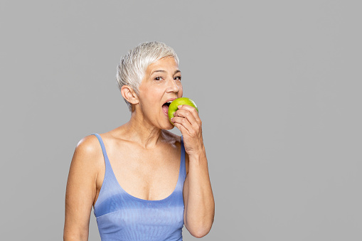 Portrait of beautiful mature woman with grey hair biting green apple. Attractive middle aged woman with beautiful smile isolated over grey background. She is looking at camera.