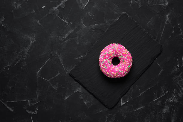istock Glazed pink donut with colored sprinkles on a black stone background. Top view, flat lay 1296013793