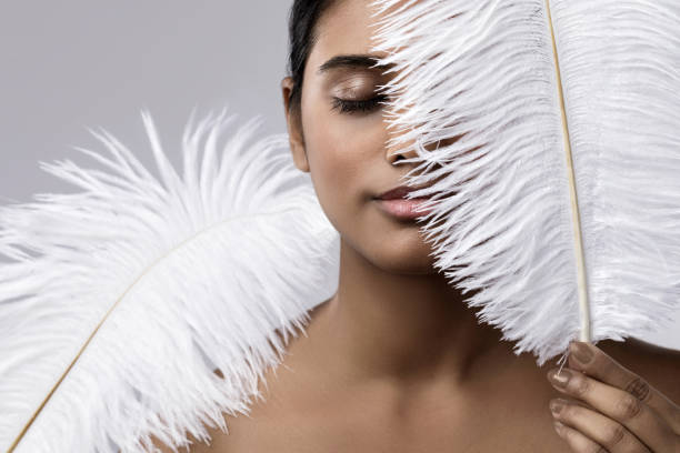 Portrait of Caucasian teenage girl wearing creative feather mask and facial decorations, standing against blue background, expressing individuality and artistic style at home