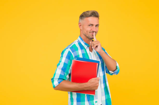 istock back to school. man tutor hold exercise book for lesson. University student on yellow background. Cheerful young man holding book and smiling. hush gesture. keep or reveal a secret. copyright 1296019378