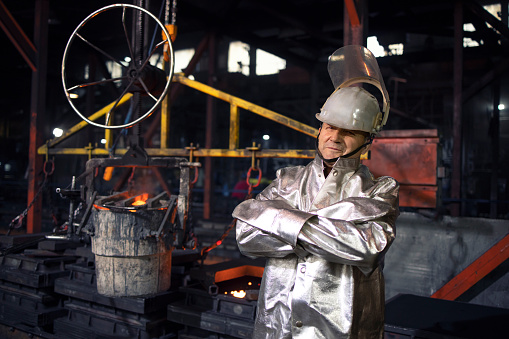 Portrait of workman in aluminized high temperature protection suit with arms crossed standing in foundry steel production factory. In background bucket with liquid molten iron. Metallurgy and heavy industry.