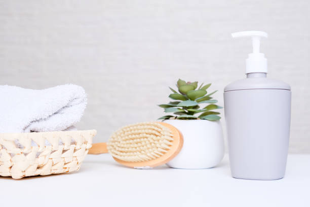istock Grey soap dispenser, spa towel, plant and massage brush on white counter table inside a bright bathroom background 1296027034
