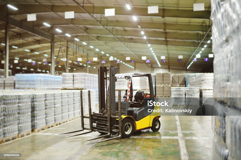 loader on the background of a huge industrial food warehouse with plastic PET bottles with beer, water, drinks. Yellow loader on the background of a huge industrial food warehouse with plastic PET bottles with beer, water, drinks. Forklift Stock Photo