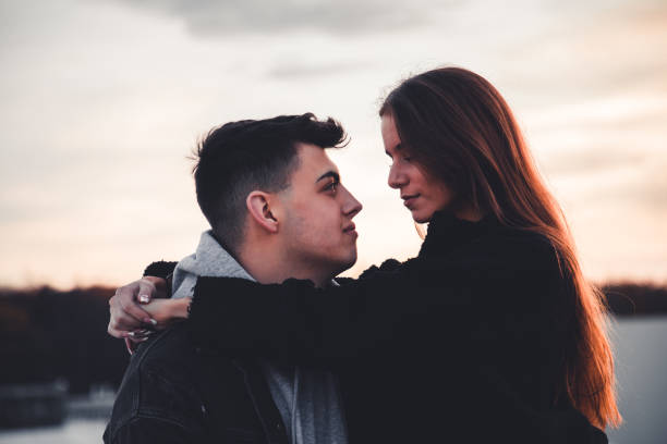 Happy millennial generation couple together in love. Embracing each other, looking into each other`s eyes under moody sunset twilight. Real People Millennial Generation Couple in Love Portrait.
