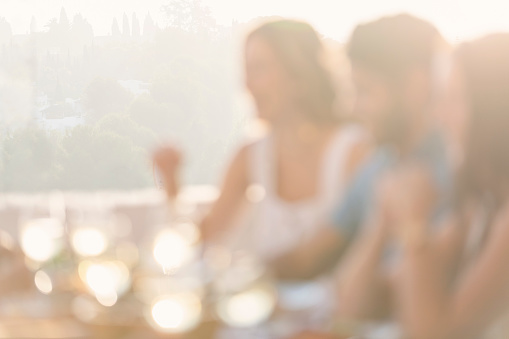 Group of friends eating a meal. Defocussed. They are drinking white wine and eating a meal and having fun. They are happy and laughing. There is wine and food on the table. Outdoors at sunset. Friendship concept with happy men and women at a restaurant. Intentionally shot out of focus for use a s a background.