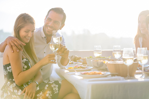 Group of friends eating a meal. They are drinking white wine and eating a meal and having fun. They are happy and laughing. There is a couple in the foreground. There is wine and food on the table. Outdoors at sunset. Friendship concept with happy men and women at a restaurant.