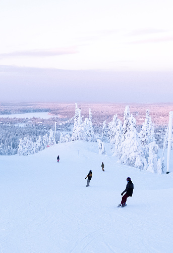 A group of young people slide down the slope on a snowboard. Landscape from the top angle. Snowy mountain, forest. Ski resort in Lapland