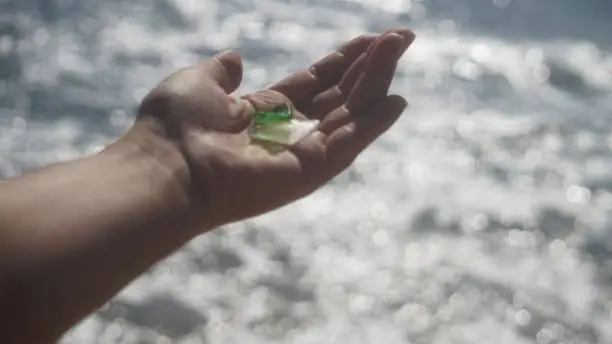 Close-up of nostalgic hand holding two bits of smooth, green and white sea-glass at the edge of the ocean, with the water sparkling in the sunlight in the background, in Sunset Beach, California.