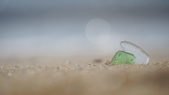 Low angle selective focus close-up of two smooth pieces of sea-glass stuck together in the sand at the back with the defocused ocean sparkling in the background.