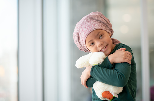 A young mixed race girl with leukaemia is holding onto her favourite teddy bear tightly. She has a headscarf covering her head as she has lost her hair in chemotherapy.