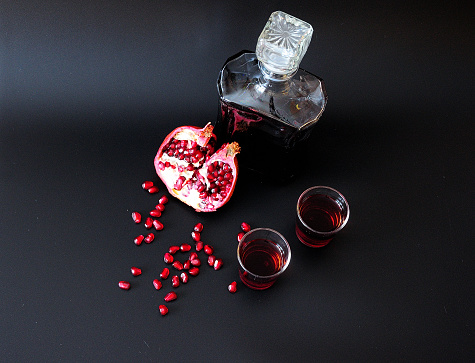 Dark red liqueur in two glasses and a glass bottle on a black matte background next to the seeds and pomegranate fruit. View from above.