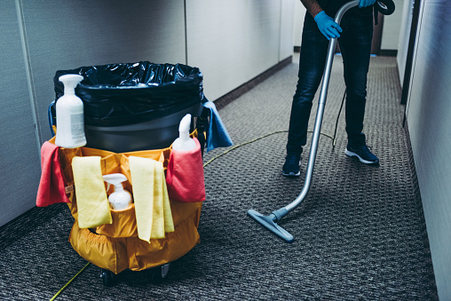 Janitor vacuum cleaning the corridor carpet floor inside an office building.around the garbage bin he has attached cleaning products and wipes.During the pandemic everything has to be disinfected