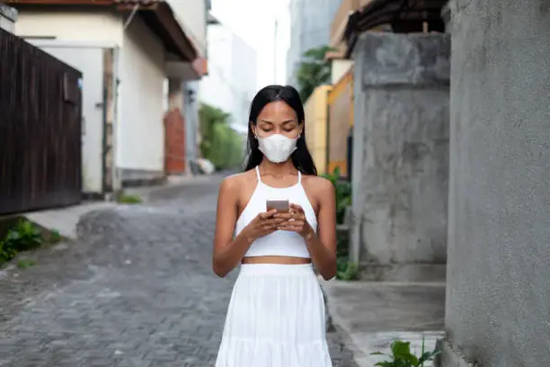 Portrait of Indonesian woman with protection face mask ,using smartphone, while walking on empty residential area