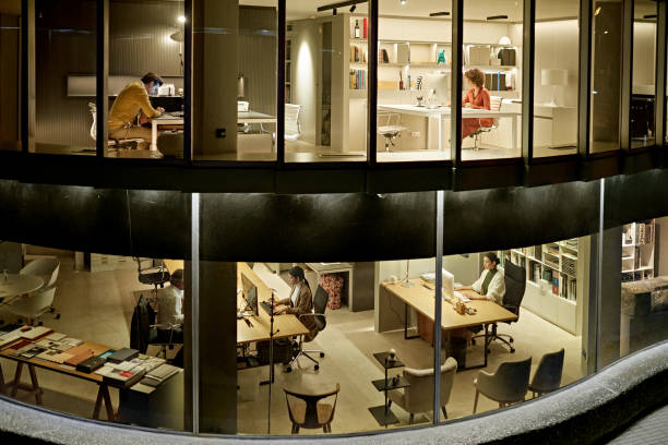 Design Professionals Working Late in Two-Story Office Personal perspective of five corporate coworkers photographed through windows sitting at their desks on two floors in modern office at night. office window night stock pictures, royalty-free photos & images