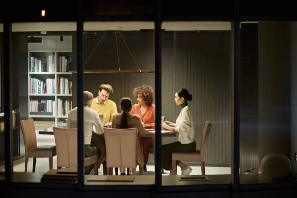 Corporate Team Working Late in Modern Office Exterior view photographed through window of male and female professionals in 20s, 30s, 40s and 50s sitting at conference table. office window night stock pictures, royalty-free photos & images