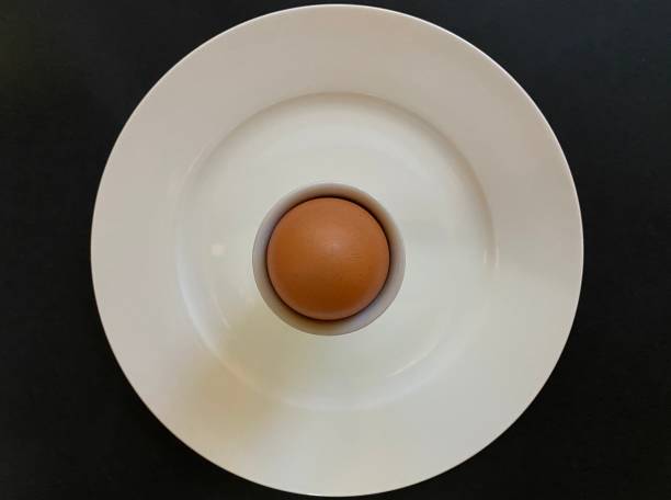 Organic Hard Boiled Egg High angle closeup view of an organic brown-shelled hard boiled egg in a white eggcup on a white ceramic plate. Black background boiled egg cut out stock pictures, royalty-free photos & images