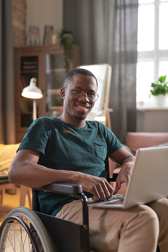 Portrait of African disabled man sitting in wheelchair with laptop on his knees and smiling at camera working at home