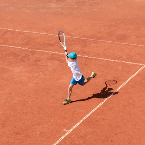 a boy plays tennis on a clay court. a little tennis player focused on the game and shot in flight after hitting the ball - tennis teenager little boys playing imagens e fotografias de stock