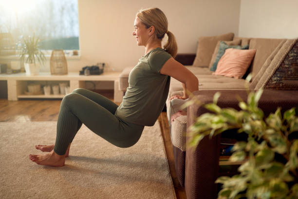 Side view of happy athlete doing triceps dips while leaning on the sofa at home. Happy sportswoman leaning on the sofa and practicing triceps dips during home workout. bicep stock pictures, royalty-free photos & images