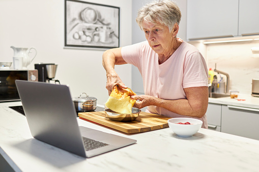 An 80-year-old senior citizen takes part in cooking classes online and uses her laptop or Macbook to cook in her own modern design kitchen