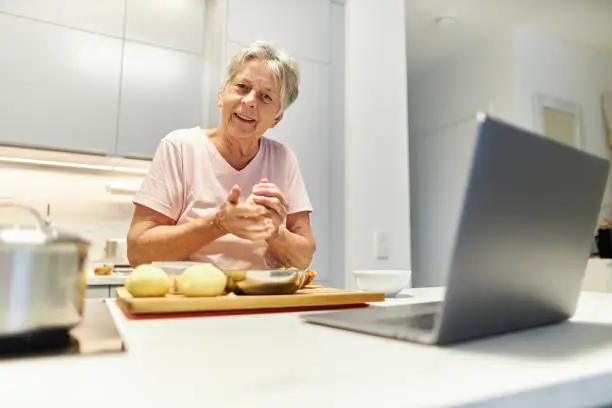 Photo of An 80-year-old senior citizen takes part in cooking classes online and uses her laptop or Macbook to cook in her own modern design kitchen