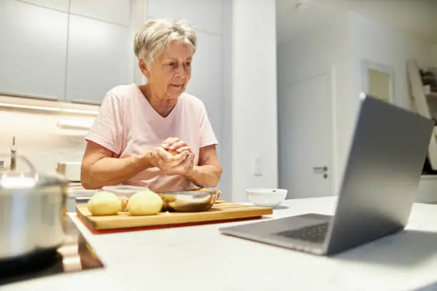 An 80-year-old senior citizen takes part in cooking classes online and uses her laptop or Macbook to cook in her own modern design kitchen, high-resolution photo with copy space
