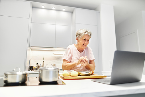 Una persona de la tercera edad de 80 años participa en clases de cocina en línea y utiliza su computadora portátil o Macbook para cocinar en su propia cocina de diseño moderno photo