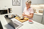 An 80-year-old senior citizen takes part in cooking classes online and uses her laptop or Macbook to cook in her own modern design kitchen