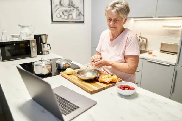 An 80-year-old senior citizen takes part in cooking classes online and uses her laptop or Macbook to cook in her own modern design kitchen, high-resolution photo with copy space