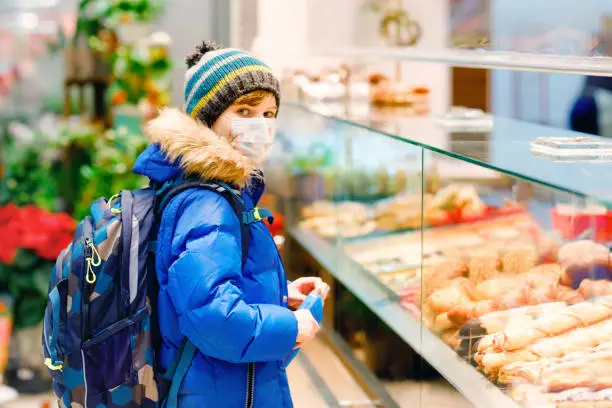 Kid boy wearing medical mask buy bread and pastry for school lunch in bakery. Child with backpack and winter clothes. Schoolkid during lockdown and quarantine time during corona pandemic disease.