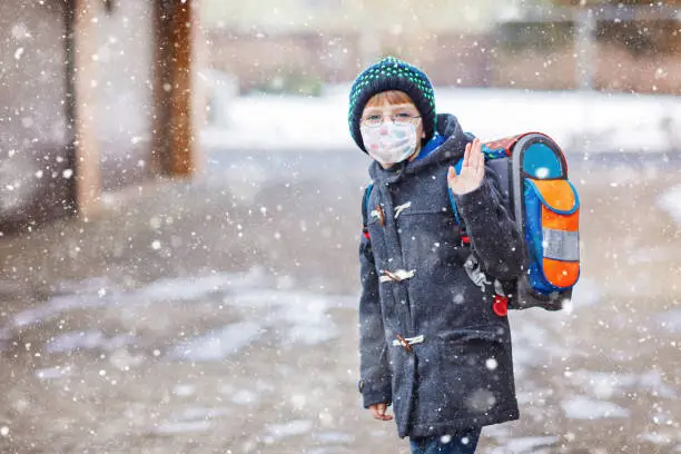 Little kid boy with glasses wearing medical mask on the way to school after lockdown. Child backpack satchel. Schoolkid on winter day with warm clothes. Quarantine time during corona pandemic disease.