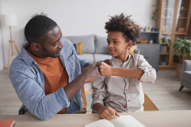 Father and Son Fistbumping Portrait of African-American father fist bumping smiling son while doing homework together at home parenting stock pictures, royalty-free photos & images