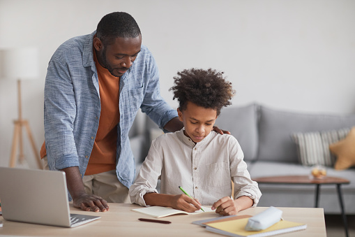 Portrait of proud African-American father helping teenage boy doing homework at desk in modern home interior, copy space