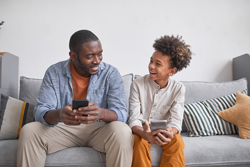Joyful teenager having fun with his father playing video game on smartphones together while staying at home, medium long shot