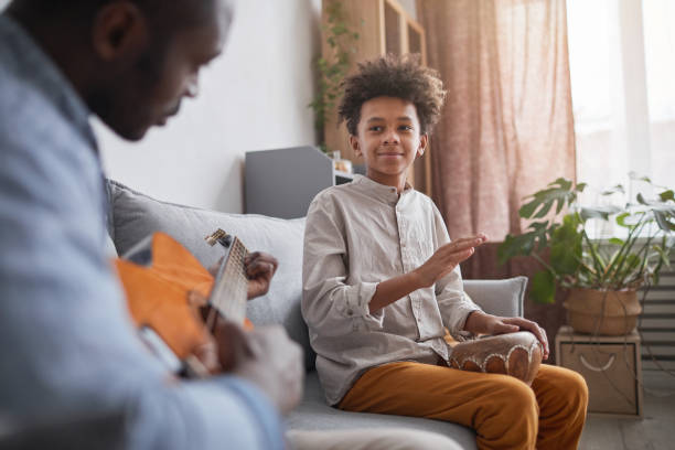 Father And Son Playing Music Mature African American man and his teen son spending great time together at home playing music on guitar and djembe father and son guitar stock pictures, royalty-free photos & images