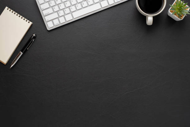 istock Top view above of Black leather office desk table with keyboard, notebook and coffee cup with equipment other office supplies. Business and finance concept. Workplace, Flat lay with blank copy space. 1296119730