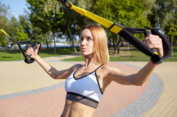 スポーツウェアで完璧な体を持つ若い運動女性は、市の公園でtrxフィットネスストラップで彼女の腕を訓練 - crossfit exercising sports training outdoors ストックフォトと画像