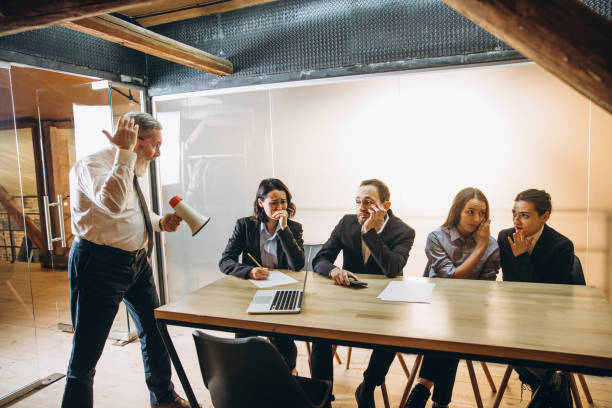istock Angry boss with megaphone screaming at employees in office, scared and annoyed colleagues listening at the table 1296127399