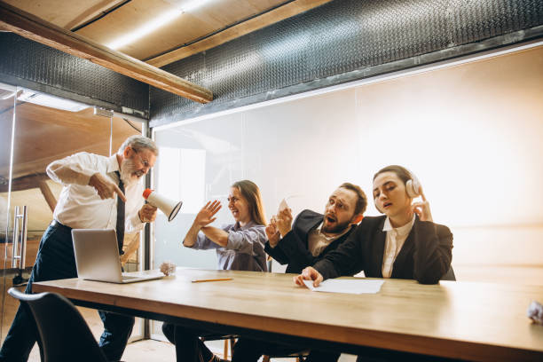 istock Angry boss with megaphone screaming at employees in office, scared and annoyed colleagues listening at the table 1296127412