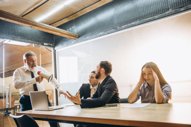 istock Angry boss with megaphone screaming at employees in office, scared and annoyed colleagues listening at the table 1296127421