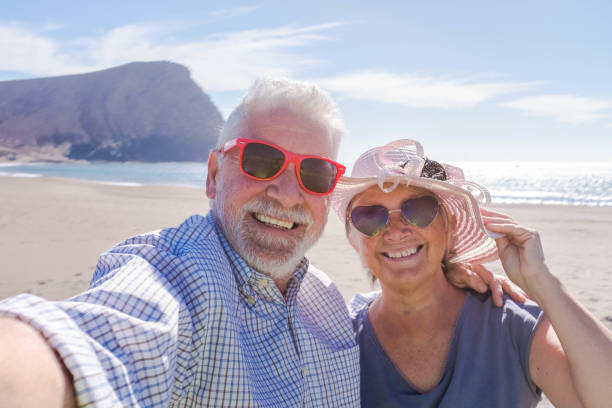 couple of seniors travelling and taking a selfie in a beautiful beach with a mountain at the background - smiling and ejoying looking at the camera wearing sun glasses - vacations lifestyle couple of seniors traveling and taking a selfie in a beautiful beach with a mountain at the background - smiling and enjoying looking at the camera wearing sun glasses - vacations lifestyle together for yes stock pictures, royalty-free photos & images