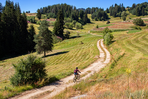 Young woman riding bike along dirt road surrounded by grass fields and flowers in a remote mountain area during summer vacation