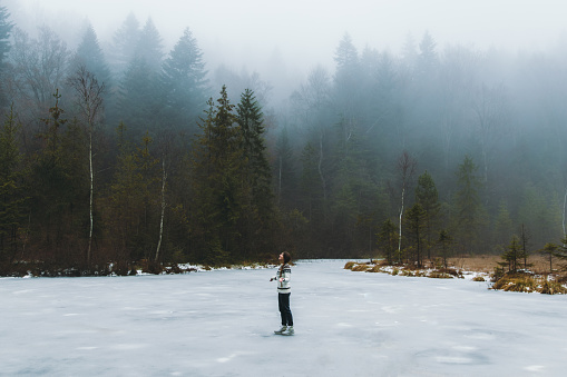 Woman with long hair, in wool sweater walking in the picturesque natural parkland looking at the beautiful icy lake and moody mountain forest