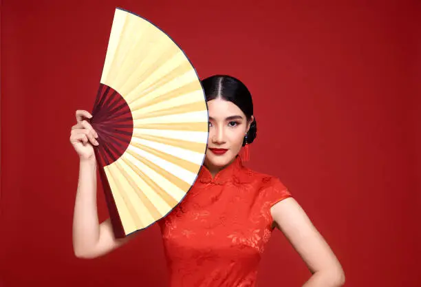 Happy Chinese new year. Asian woman wearing traditional cheongsam qipao dress holding fan isolated on red background.