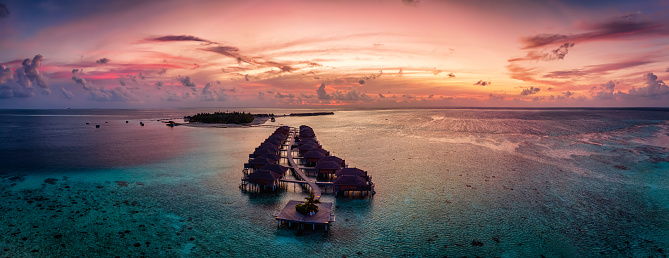 Aerial panorama of a tropical paradise island in the Maldives, Indian Ocean, with water lodges over the turquoise reef during sunset time