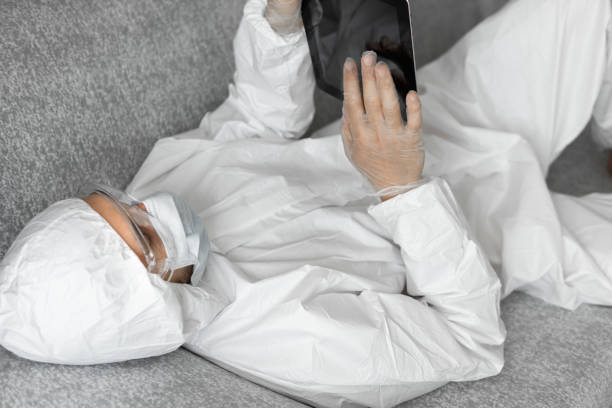 istock Man in protective white suit and medical mask is working from home lying on a sofa with a tablet because of coronavirus epidemic. Remote work during pandemic. Stay home during COVID-19 quarantine concept. 1296151299