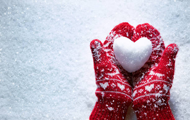 manos femeninas en manoplas de punto con corazón nevado contra fondo de nieve - holidays fotografías e imágenes de stock